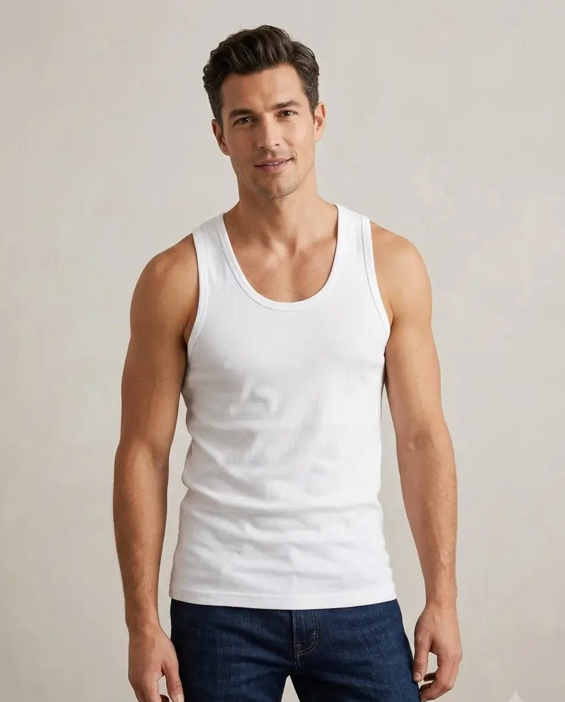 A man wearing a white ribbed tank top and dark blue jeans, standing against a light beige studio background.