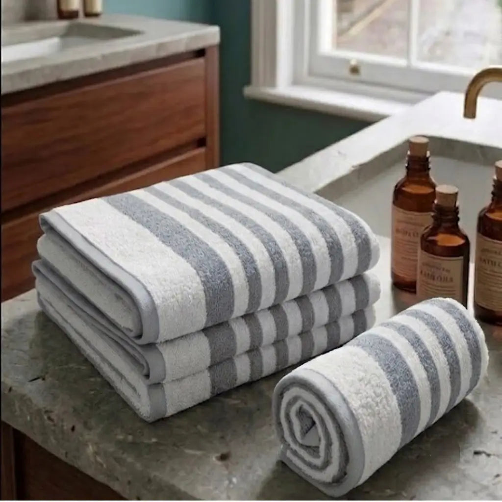 A set of three folded and rolled bath towels with gray and white stripes, displayed on a gray stone bathroom counter next to amber bottles.