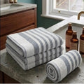 A set of three folded and rolled bath towels with gray and white stripes, displayed on a gray stone bathroom counter next to amber bottles.