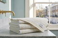 A stack of folded white towels with a small 'ALAM' label sits on a marble countertop in a bathroom setting with a gold faucet and a window in the background.
